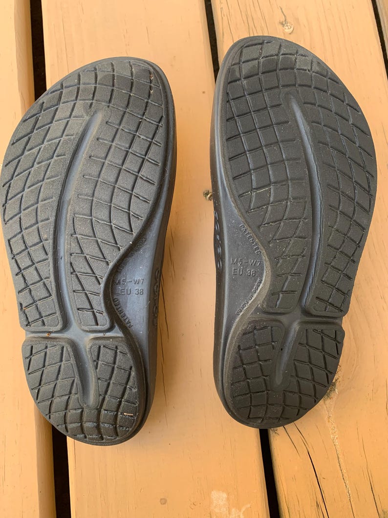 May include: A pair of black sandals, viewed from the soles. The sandals have a textured, grid-like pattern and a contoured design. The size is M5-W7, EU 38. The sandals are on a wooden surface.