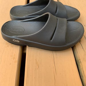 May include: A pair of black slide sandals with a contoured footbed and two straps across the top. The sandals are made of a rubber-like material and are designed for comfort. The brand name "OOFOS" is visible on the footbed.