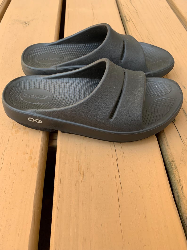 May include: A pair of black slide sandals with a contoured footbed and two straps across the top. The sandals are made of a rubber-like material and are designed for comfort. The brand name "OOFOS" is visible on the footbed.