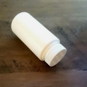 May include: A white, cylindrical container with a perforated cap. The container is made of a smooth, opaque material and is resting on a dark wooden surface. The cap has small holes for dispensing the contents.