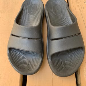 May include: A pair of black slide sandals. The sandals have a textured footbed and two straps across the top of the foot. The brand name is visible on the footbed. The sandals are made of a rubber-like material.
