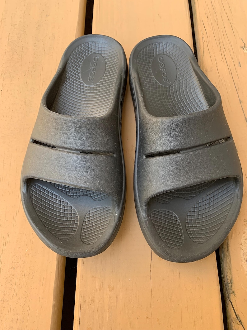 May include: A pair of black slide sandals. The sandals have a textured footbed and two straps across the top of the foot. The brand name is visible on the footbed. The sandals are made of a rubber-like material.