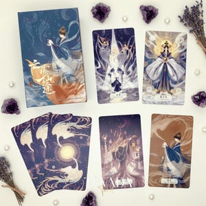 May include: A tarot card deck with a blue and orange box featuring a woman in flowing robes. Several tarot cards are displayed, each with unique illustrations of figures in elegant attire. Amethyst crystals and lavender sprigs surround the cards.