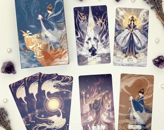 Puede incluir: Una baraja de cartas del tarot con una caja azul y naranja que presenta a una mujer con túnicas fluidas. Se muestran varias cartas del tarot, cada una con ilustraciones únicas de figuras con atuendos elegantes. Cristales de amatista y ramitas de lavanda rodean las cartas.
