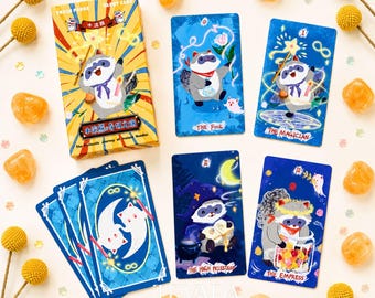 May include: A tarot card deck featuring raccoon illustrations, including a box labeled Trash Panda Tarot Card and The Fantastic Journey of Instant Noodles, with cards titled The Fool, The Magician, The High Priestess, and The Empress.