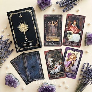 Könnte beinhalten: Eine schwarze Tarotkartenbox mit goldenen Akzenten und der Aufschrift "Starry Night". Mehrere Tarotkarten werden gezeigt, mit Illustrationen und Titeln wie "The High Priestess" und "The Lovers". Amethystkristalle und Lavendelzweige umgeben die Karten.