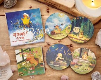 May include: A collection of Cleo Duck Tarot cards and coasters. The tarot card box is square with a blue background and a yellow duck illustration. The coasters feature duck illustrations in various scenes. Text in Chinese and English is visible on the box and coasters.