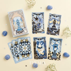 May include: A light blue tarot card deck with the words "Chess Tarot" on the box. The cards feature illustrations of chess pieces and rabbits in a whimsical style, with blue and black accents. Decorative blue stones and flowers surround the deck.