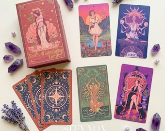 Op de afbeelding: Een tarotkaartspel getiteld "She's Wilderness Tarot" met verschillende kaarten afgebeeld. De kaarten bevatten illustraties van figuren en symbolen in verschillende kleuren. De doos is rood met gouden accenten. Amethistkristallen en lavendel takjes omringen de kaarten.