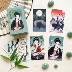 Puede incluir: Un juego de cartas de tarot que incluye una caja azul claro con la ilustración de una persona escribiendo, junto a cuatro cartas individuales que muestran escenas con figuras en vestimenta tradicional, luna llena, un puente de piedra y bambú, más piedras verdes.