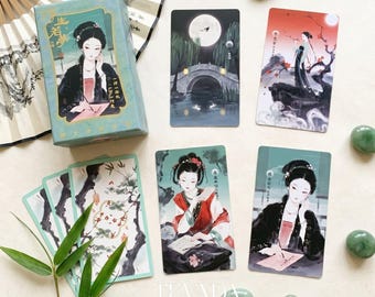 May include: A tarot card deck set featuring a light blue box with an illustration of a person writing, alongside four individual cards depicting scenes with figures in traditional attire, a full moon, a stone bridge, and bamboo, plus green stones.
