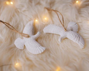 May include: Two white dove ornaments with textured wings and attached twine hangers. The ornaments are lying on a soft, cream-colored surface, with warm fairy lights in the background. These decorative ornaments are ideal for holiday decor.