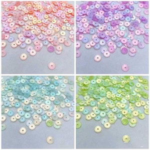 May include: A collection of small, round, flat sequins with a central hole and a radial ribbed texture. The sequins are organized into four distinct color groups: peach and pink, purple and lavender, light blue and teal, and lime green and yellow.