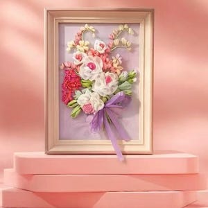 Puede incluir: Una obra de arte floral enmarcada con un ramo de flores blancas, rosas y rojas con hojas verdes y una cinta morada. La obra se exhibe sobre una plataforma rosa, sobre un fondo rosa.