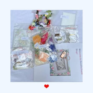 K&ouml;nnte beinhalten: Eine Sammlung von Bastelmaterialien, darunter Stickgarn, B&auml;nder, Stoff und Perlen, verpackt in transparenten Plastikt&uuml;ten. Ein gerahmtes Blumendesign und der Text "Welcome to Needlecharm Shop!" sind ebenfalls zu sehen.