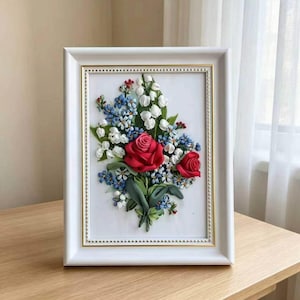 May include: A framed floral artwork featuring a bouquet of red roses, white lilies, and blue forget-me-nots. The flowers are arranged on a white background within a white frame with gold accents. The artwork is displayed on a wooden table.