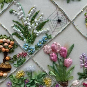 K&ouml;nnte beinhalten: Gestickte Blumenkunst mit verschiedenen Blumen, darunter wei&szlig;es Maigl&ouml;ckchen, rosa Tulpen und blaue Vergissmeinnicht. Das Design umfasst einen kleinen Baum mit orangen Fr&uuml;chten und ein Spinnennetz. Das Kunstwerk ist mit silbernen Zierleisten gerahmt.