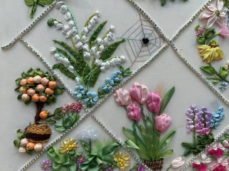 K&ouml;nnte beinhalten: Gestickte Blumenkunst mit verschiedenen Blumen, darunter wei&szlig;es Maigl&ouml;ckchen, rosa Tulpen und blaue Vergissmeinnicht. Das Design umfasst einen kleinen Baum mit orangen Fr&uuml;chten und ein Spinnennetz. Das Kunstwerk ist mit silbernen Zierleisten gerahmt.