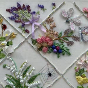 K&ouml;nnte beinhalten: Eine wei&szlig;e dekorative Tafel mit gestickten Blumendesigns. Die Tafel zeigt Lavendel, Rosen, Calla-Lilien und andere Blumen sowie eine Libelle, einen Schmetterling und ein Spinnennetz. Die Gitterlinien sind mit funkelnden Strasssteinen akzentuiert.