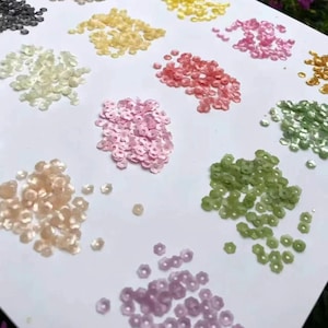 Op de afbeelding: Een verzameling kleine, bloemvormige lovertjes gerangschikt in afzonderlijke hoopjes op een wit oppervlak. De lovertjes zijn verkrijgbaar in verschillende kleuren, waaronder blauw, geel, roze, groen, paars, wit, perzik en zwart, elk met een gaatje.