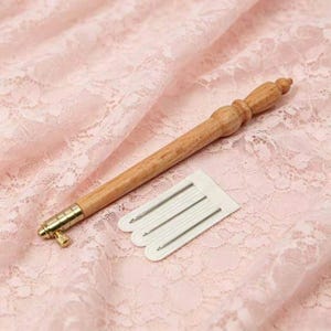 May include: A wooden punch needle tool with a light brown finish and a gold-toned metal tip, accompanied by a white plastic needle threader containing three thin, silver-coloured metal needles arranged in a parallel row.