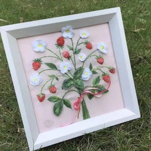 May include: A framed embroidery of strawberries and white flowers. The artwork features red strawberries, white flowers with yellow centers, and green leaves, all tied together with a pink ribbon. The embroidery is set against a pink background and framed in a white wooden frame.