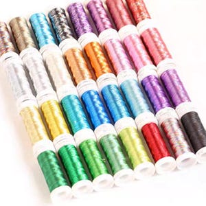 May include: A collection of twenty-eight cylindrical spools of metallic embroidery thread arranged in a grid. The spools feature a variety of vibrant colors including green, blue, yellow, red, pink, purple, silver, gold, and copper tones.