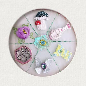 May include: A circular, embroidered artwork featuring various whimsical elements. Includes a purple flower, a mushroom, a bottle, a geometric shape, a butterfly, and a blue and yellow shape. The design is set against a white background with green stitching.