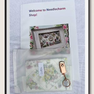 K&ouml;nnte beinhalten: Ein gerahmter Druck mit floraler Stickerei, eine transparente Plastikt&uuml;te mit einem Muster und ein ros&eacute;goldener Schl&uuml;sselanh&auml;nger. Der Druck ist von rosa Rosen umgeben. Der Text "Welcome to Needlecharm Shop!" befindet sich oben im Bild.