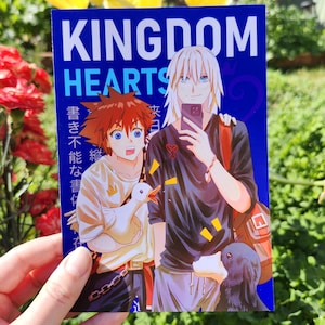 May include: A blue and white Kingdom Hearts themed card featuring two anime characters. The character on the left has red hair and is holding a white duck. The character on the right has white hair and is holding a phone. The text on the card reads "Kingdom Hearts" in large white letters and "書ききれない言葉" in smaller black letters.