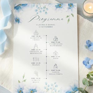 Può includere: Invito di nozze con un design floreale blu. L'invito include i nomi Claudia e Daniel, la data e un programma degli eventi. Sono visibili anche un anello d'oro, una candela e un nastro blu.