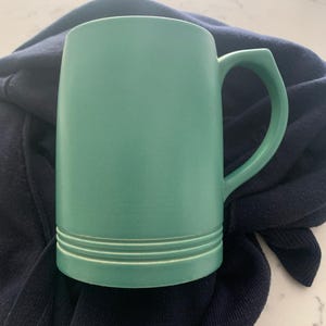 May include: A green ceramic beer stein with a handle. The stein has two raised lines around the bottom.