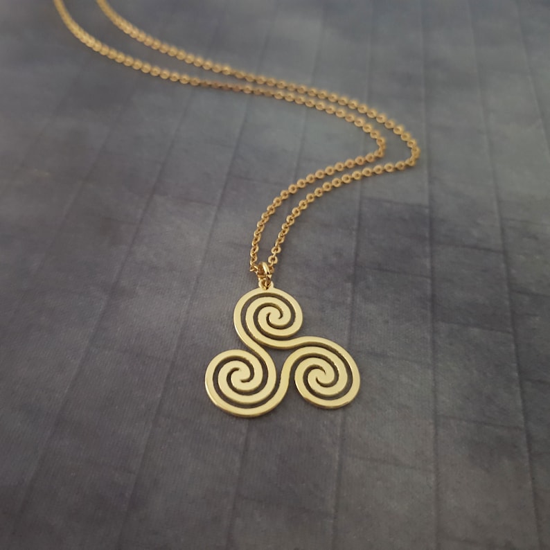 K&ouml;nnte beinhalten: Goldfarbene Anh&auml;ngerkette mit einem keltischen Triskele-Design. Der Anh&auml;nger hat drei miteinander verbundene Spiralen. Die Halskette ist filigran und goldfarben, vor grauem Hintergrund.