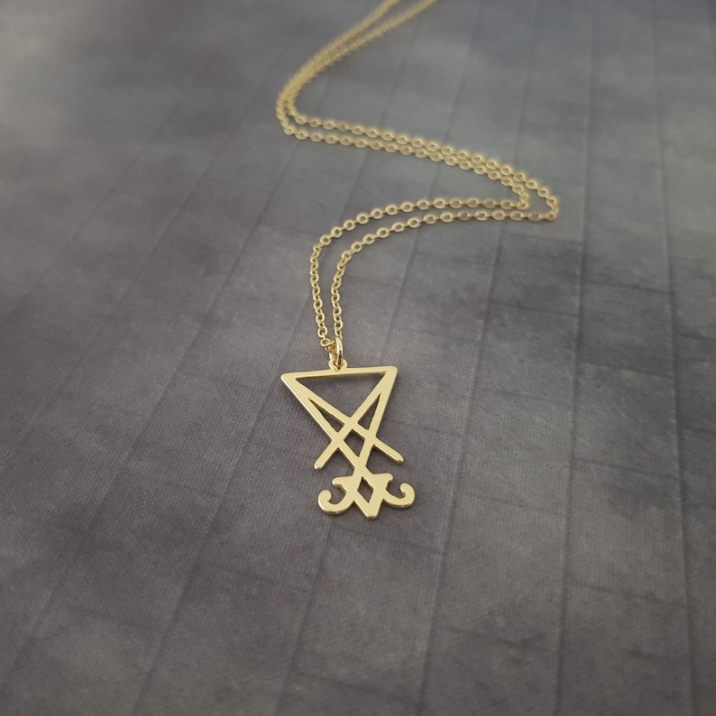 May include: A gold chain necklace with a pendant featuring the sigil of the demon Baphomet. The sigil is a stylized inverted pentagram with two horns at the bottom.