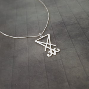 May include: A silver necklace with a pendant featuring a stylized sigil, a triangular design with intersecting lines and a symbol at the bottom.