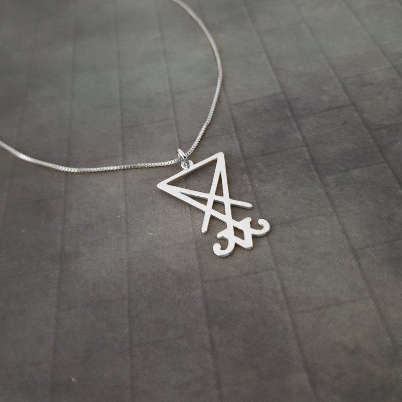 May include: A silver necklace with a pendant featuring a stylized sigil, a triangular design with intersecting lines and a symbol at the bottom.