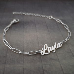 Può includere: Bracciale color argento con il nome "Layla" in corsivo. Il bracciale ha un design a catena, lunghezza regolabile e chiusura. Il gioiello è esposto su una superficie grigia.