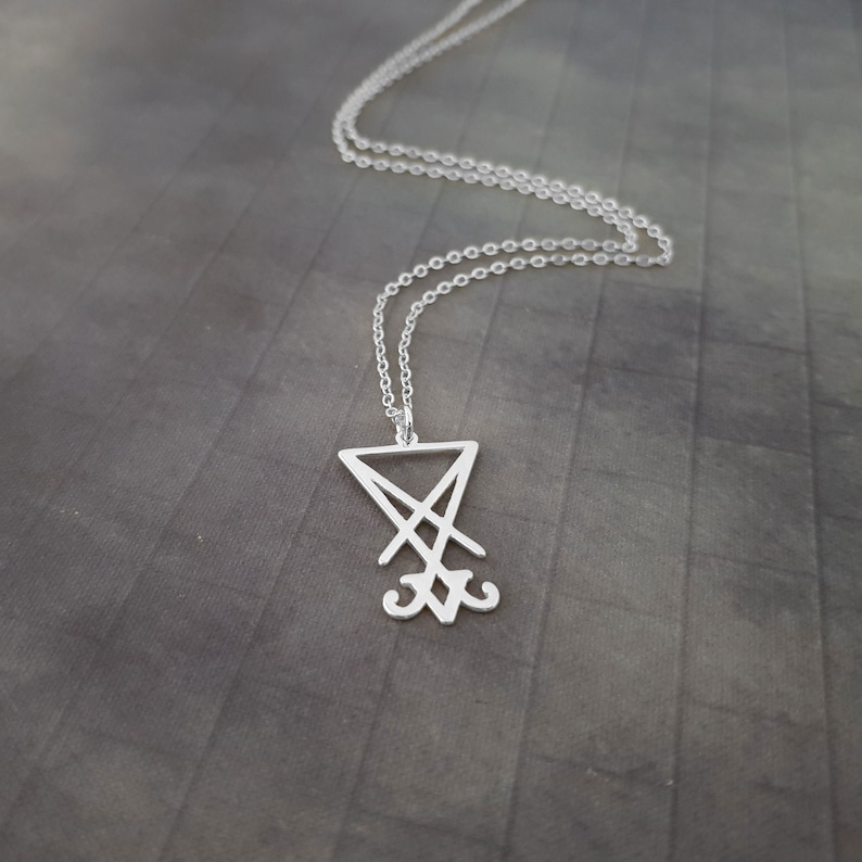 May include: A silver necklace with a pendant featuring the sigil of the demon Baphomet. The pendant is a triangular shape with a cross inside and two horns at the bottom.