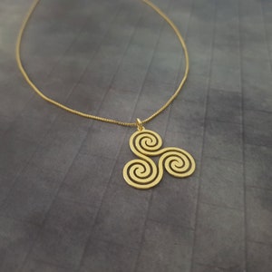 K&ouml;nnte beinhalten: Eine goldfarbene Halskette mit Anh&auml;nger im keltischen Triskele-Design. Der Anh&auml;nger hat drei miteinander verbundene Spiralen. Die Halskette ist d&uuml;nn und goldfarben, vor grauem Hintergrund.