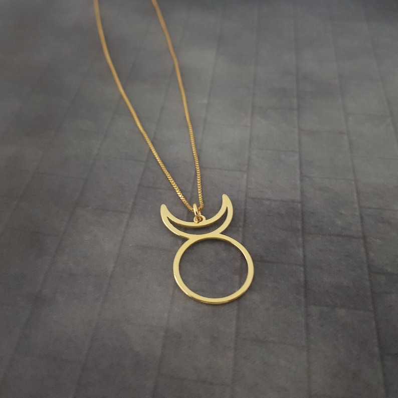 Op de afbeelding: Een gouden ketting met een hanger met een cirkel met twee halve manen erop. De hanger is een eenvoudig, minimalistisch ontwerp.