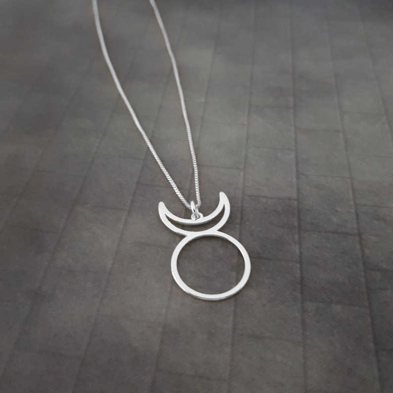 Op de afbeelding: Een zilveren ketting met een hanger in de vorm van een cirkel met twee halve manen erboven. De hanger heeft een eenvoudig, minimalistisch ontwerp.