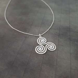 K&ouml;nnte beinhalten: Eine silberfarbene Halskette mit einem Anh&auml;nger im keltischen Triskele-Design. Der Anh&auml;nger, mit drei miteinander verbundenen Spiralen, h&auml;ngt an einer feinen Kette. Die Halskette wird vor einem dunklen, strukturierten Hintergrund gezeigt.