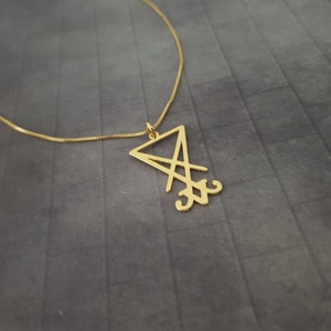 May include: A gold chain necklace with a pendant featuring a stylized sigil in the shape of a triangle with a cross inside.