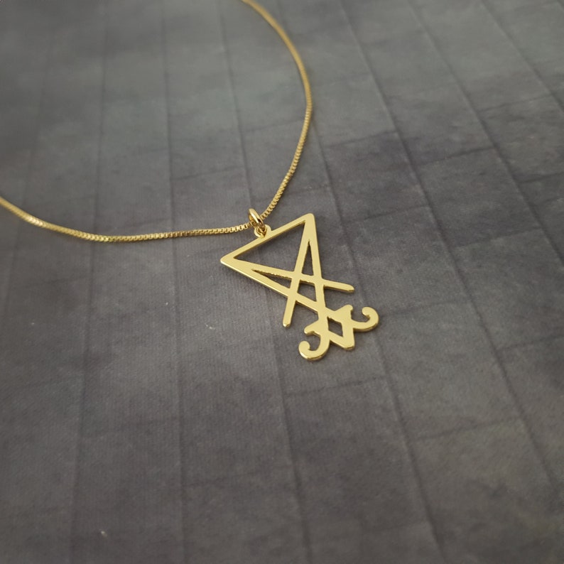 May include: A gold chain necklace with a pendant featuring a stylized sigil in the shape of a triangle with a cross inside.