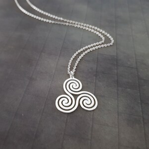 K&ouml;nnte beinhalten: Eine silberfarbene Halskette mit einem keltischen Triskele-Design. Der Anh&auml;nger, ein Symbol aus drei Spiralen, h&auml;ngt an einer feinen Kette. Die Halskette wird vor einem dunkelgrauen Hintergrund gezeigt.