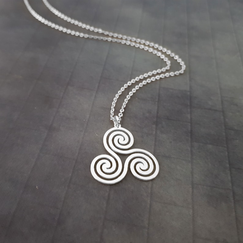 K&ouml;nnte beinhalten: Eine silberfarbene Halskette mit einem keltischen Triskele-Design. Der Anh&auml;nger, ein Symbol aus drei Spiralen, h&auml;ngt an einer feinen Kette. Die Halskette wird vor einem dunkelgrauen Hintergrund gezeigt.