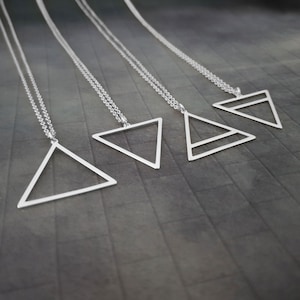 May include: Four silver-colored geometric triangle necklaces. Each necklace features a different open triangle design suspended from a delicate chain. The necklaces are displayed against a dark gray background.