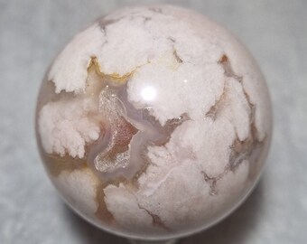 May include: A white agate sphere with brown and pink markings. The sphere is polished and has a smooth surface.