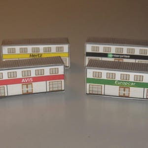 May include: Four miniature building models, each representing a car rental company: Hertz, Avis, Enterprise, and Europcar. The buildings have white walls, brown roofs, and colored stripes with the company names.