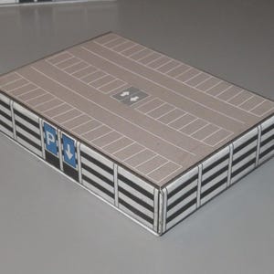 May include: A miniature parking garage model with a tan roof featuring parking space markings and directional arrows. The sides are detailed with black and white striped panels and blue parking signs.
